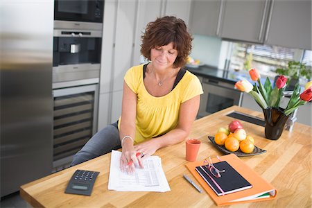 simsearch:6102-04929446,k - Woman sitting in kitchen doing paperwork, smiling Stock Photo - Premium Royalty-Free, Code: 6102-04929449