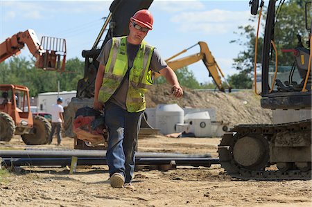 simsearch:6105-07744367,k - Construction engineer carrying pipe cutting saw at heavy construction site with excavator Foto de stock - Sin royalties Premium, Código: 6105-07744373