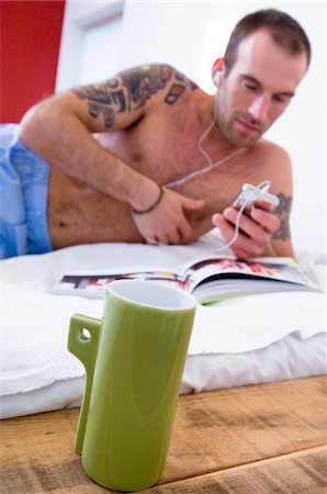 simsearch:6108-05856541,k - Tattooed man, barechested, listening to iPod player, lying on a bed, mug in foreground Fotografie stock - Premium Royalty-Free, Codice: 6108-05856532