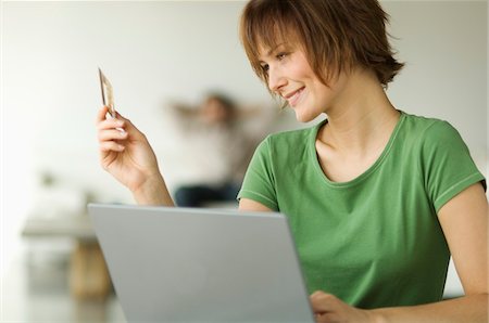 Smiling woman using laptop computer, holding credit card, man in the background Stock Photo - Premium Royalty-Free, Code: 6108-05856725