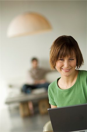 Smiling woman using laptop computer, man in the background Stock Photo - Premium Royalty-Free, Code: 6108-05856726