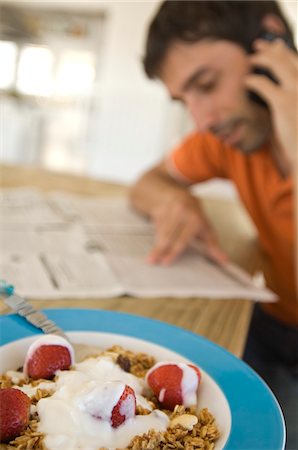 reading and eating - Young man reading newspaper, using mobile phone, strawberries and cereals in foreground Stock Photo - Premium Royalty-Free, Code: 6108-05858523