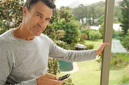 Man leaning against bay window, holding mobile phone Stock Photo - Premium Royalty-Free, Code: 6108-05858652