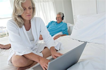 Woman using laptop in bedroom, man with headphones in background Stock Photo - Premium Royalty-Free, Code: 6108-05858703