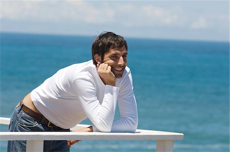 simsearch:6108-05856096,k - Young smiling man leaning against balustrade, sea in background Fotografie stock - Premium Royalty-Free, Codice: 6108-05859065