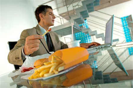 Mid adult businessman eating French fries, using laptop Foto de stock - Royalty Free Premium, Número: 6108-05859380