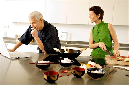 Mature man using a laptop with a mid adult woman chopping vegetables in the kitchen Foto de stock - Sin royalties Premium, Código: 6108-05859979