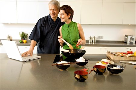 Mid adult woman preparing food with a mature man using a laptop in the kitchen Foto de stock - Sin royalties Premium, Código: 6108-05859989