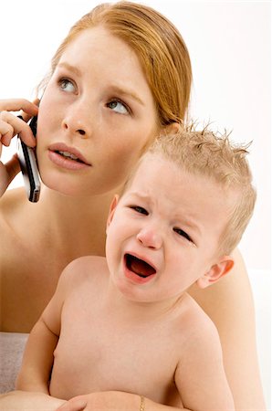 Close-up of a young woman talking on a mobile phone and holding a crying baby boy Stock Photo - Premium Royalty-Free, Code: 6108-05860152