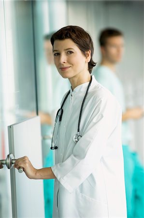 simsearch:6108-05860406,k - Portrait of a female doctor holding a door handle and smiling Fotografie stock - Premium Royalty-Free, Codice: 6108-05860305