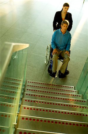 Male patient sitting in a wheelchair and a mid adult woman standing beside him near a staircase Stock Photo - Premium Royalty-Free, Code: 6108-05860407