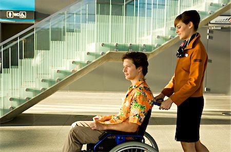 Side profile of a female cabin crew pushing a mid adult man sitting in a wheelchair Stock Photo - Premium Royalty-Free, Code: 6108-05860413