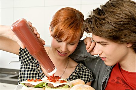 Young woman pouring tomato sauce on burgers with a teenage boy standing beside her Foto de stock - Royalty Free Premium, Número: 6108-05861231