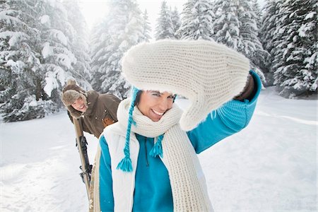 simsearch:6108-05867016,k - Young woman in winter clothes smiling at camera, man holding skis in background Fotografie stock - Premium Royalty-Free, Codice: 6108-05867041