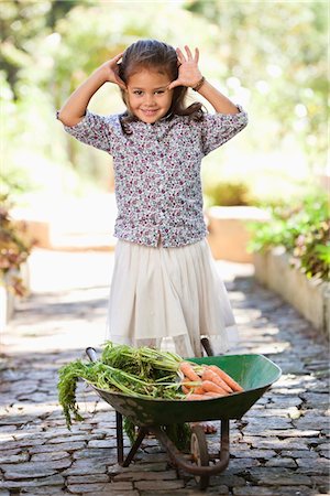 Cute girl standing near a wheelbarrow filled with carrots Stock Photo - Premium Royalty-Free, Code: 6108-05869498