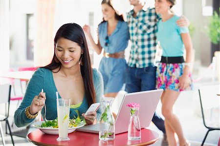 simsearch:6108-06905858,k - Smiling woman eating food while using mobile with people in the background at a restaurant Foto de stock - Sin royalties Premium, Código: 6108-05869848