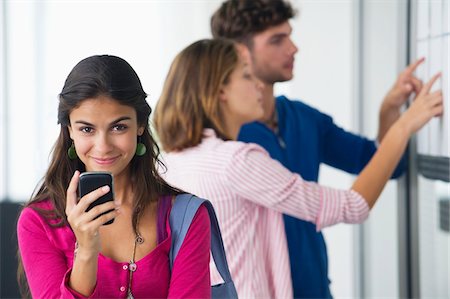 people looking at notice board - Portrait of university students using a mobile phone while friends checking for test results in bulletin board Foto de stock - Sin royalties Premium, Código: 6108-05872280