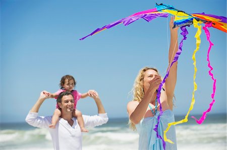 Little girl sitting on father's shoulder while mother flying shaped kite on the beach Foto de stock - Sin royalties Premium, Código: 6108-05872533