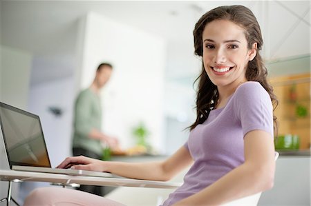 side view of someone sitting in chair - Woman using a laptop with her husband preparing food in the background Stock Photo - Premium Royalty-Free, Code: 6108-06166447