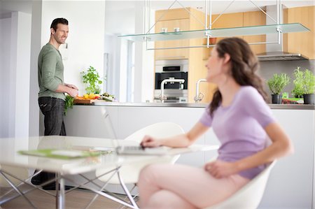 spouses laptop - Woman using a laptop with her husband preparing food in the background Stock Photo - Premium Royalty-Free, Code: 6108-06166451