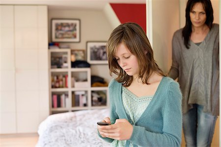 Girl text messaging on mobile phone with her mother watching her from behind the door Stock Photo - Premium Royalty-Free, Code: 6108-06167221
