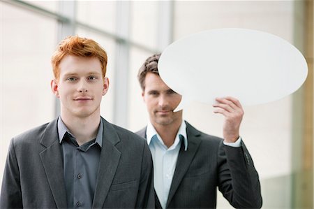 Portrait of a businesswoman smiling with her colleague holding speech bubble behind her Stock Photo - Premium Royalty-Free, Code: 6108-06167903