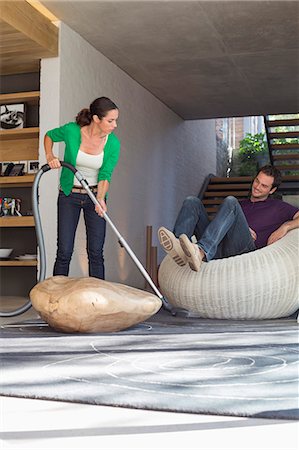 Woman cleaning house with a vacuum cleaner with her husband sitting on a seat Stock Photo - Premium Royalty-Free, Code: 6108-06906432