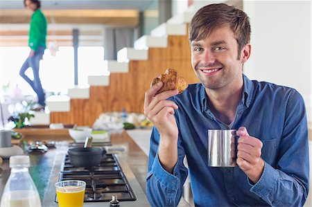 Man having breakfast at a kitchen counter with his wife in the background Stock Photo - Premium Royalty-Free, Code: 6108-06908086