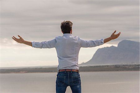 Rear view of a man standing at lakeshore with his arm outstretched Stock Photo - Premium Royalty-Free, Code: 6108-08662938