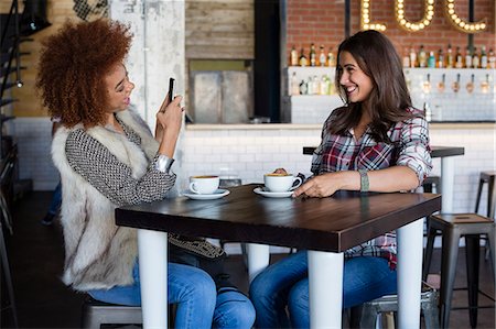 simsearch:632-08331610,k - Young woman taking a picture of her friend with smartphone at cafe Stock Photo - Premium Royalty-Free, Code: 6108-08725374