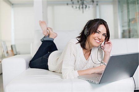 simsearch:6108-05862347,k - Beautiful brunette woman on a couch with computer and headphones smiling at camera Photographie de stock - Premium Libres de Droits, Code: 6108-08909143