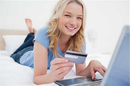 Une femme allongée sur son lit, avec son ordinateur portable et carte de crédit, les éléments de commande et souriant. Photographie de stock - Premium Libres de Droits, Code: 6109-06003027