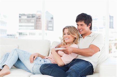 A man changes the channel while embracing a woman as they sit on the couch smiling. Stock Photo - Premium Royalty-Free, Code: 6109-06003129