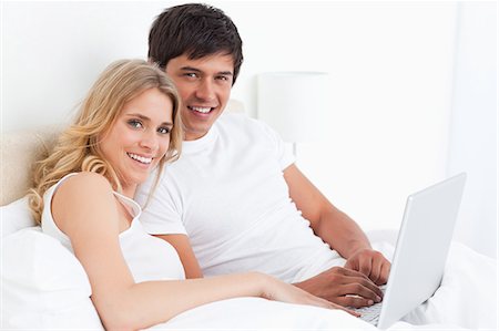 simsearch:6109-06003220,k - Un homme et une femme avec un ordinateur portable sur le lit sourient comme ils regardent en face d'eux. Photographie de stock - Premium Libres de Droits, Code: 6109-06003263