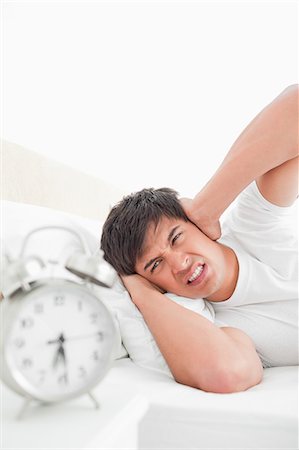 The man is covering his ears to soften the sound of the loud ringing alarm clock while in bed. Stock Photo - Premium Royalty-Free, Code: 6109-06003297