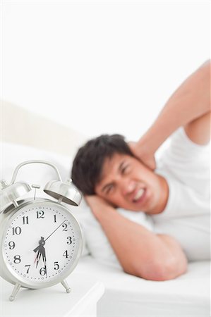 The focus is on the ringing alarm clock as the man in the background covers his ears in anger. Stock Photo - Premium Royalty-Free, Code: 6109-06003296