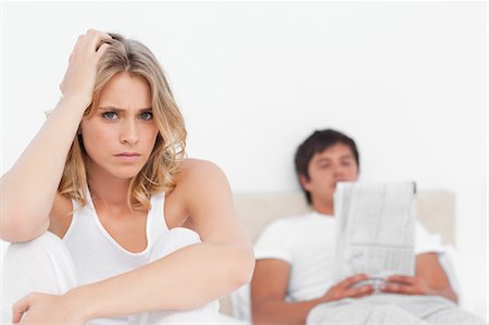 relationship distraught - The woman sits at the edge looking upset while the man sits at the back of the bed reading his paper. Stock Photo - Premium Royalty-Free, Code: 6109-06003236