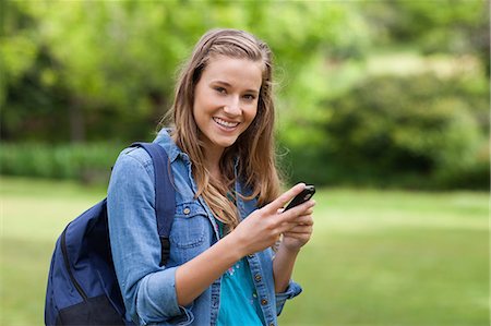Young smiling girl sending a text with her cellphone while standing up in a park Stock Photo - Premium Royalty-Free, Code: 6109-06003386