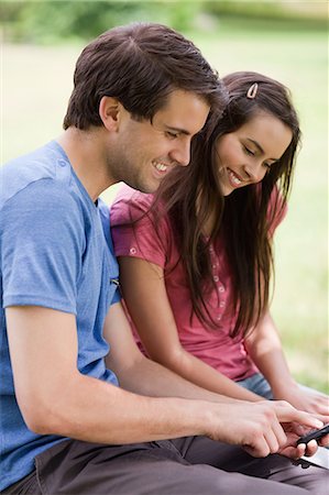 phone white woman young - Young smiling man showing his cellphone to his girlfriend while sitting in a parkland Stock Photo - Premium Royalty-Free, Code: 6109-06003449