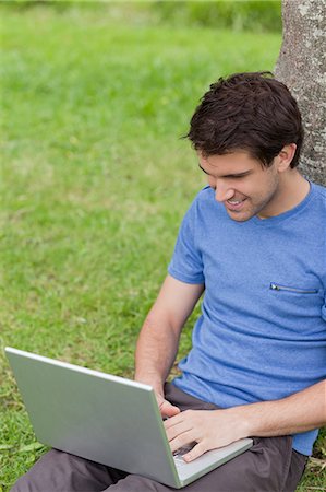 Young smiling man working on his laptop while leaning against a tree in a parkland Stock Photo - Premium Royalty-Free, Code: 6109-06003489