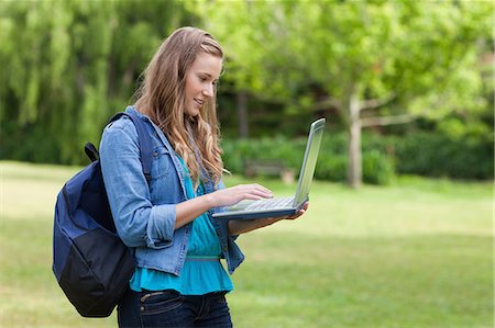 simsearch:6109-06003402,k - Young girl typing on her laptop while standing in a park and carrying a backpack Foto de stock - Sin royalties Premium, Código: 6109-06003408