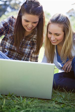 simsearch:6109-06003537,k - Young women sitting on the grass in a parkland while smiling by looking at a laptop Fotografie stock - Premium Royalty-Free, Codice: 6109-06003529