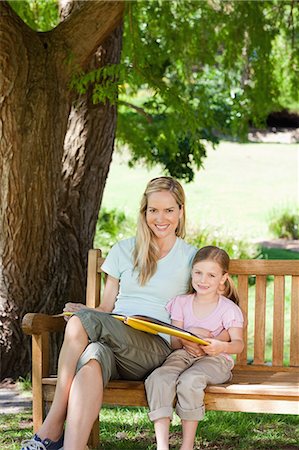 simsearch:6109-06003988,k - A smiling mom and her daughter smile and look at the camera with a book in hand and sitting on the park bench Foto de stock - Sin royalties Premium, Código: 6109-06003947