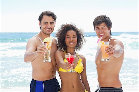 simsearch:6109-06004181,k - Two men and a woman in swimsuits smiling as they offer cocktails on a beach Foto de stock - Royalty Free Premium, Número: 6109-06004183