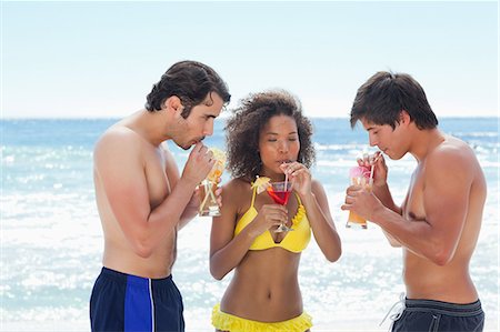 simsearch:6109-06004181,k - Two men and a woman in swimsuits drinking cocktails as they stand on a beach Foto de stock - Royalty Free Premium, Número: 6109-06004184