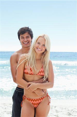 embrace couple swim suit - Woman in swimwear smiling as her friends arms are wrapped around her while they stand bt the water Stock Photo - Premium Royalty-Free, Code: 6109-06004190