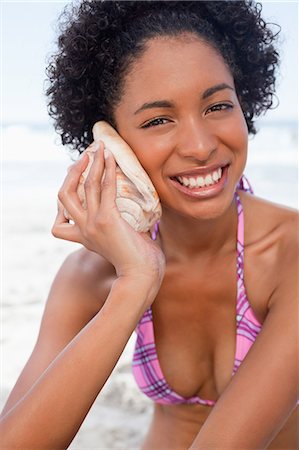 Young smiling woman in beachwear looking at the camera while listening to a shell Stock Photo - Premium Royalty-Free, Code: 6109-06004121