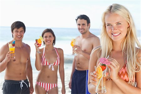 female swimsuit with friends - Woman in a bikini enjoying a cocktail on the beach while two men and a woman stand behind her Stock Photo - Premium Royalty-Free, Code: 6109-06004265