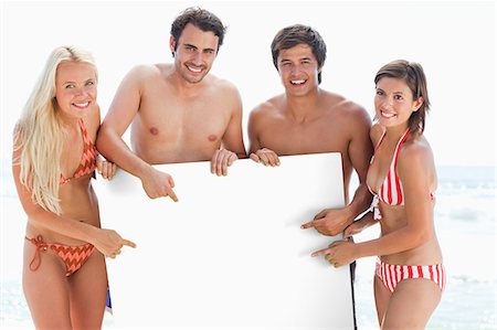 Two men and two women smiling and pointing at a large blank poster which they hold between them Stock Photo - Premium Royalty-Free, Code: 6109-06004238