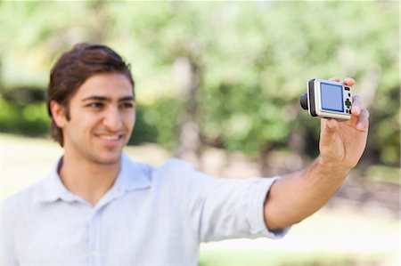 person holding a camera - Side view of a smiling young man in the park taking a picture of himself Stock Photo - Premium Royalty-Free, Code: 6109-06004497